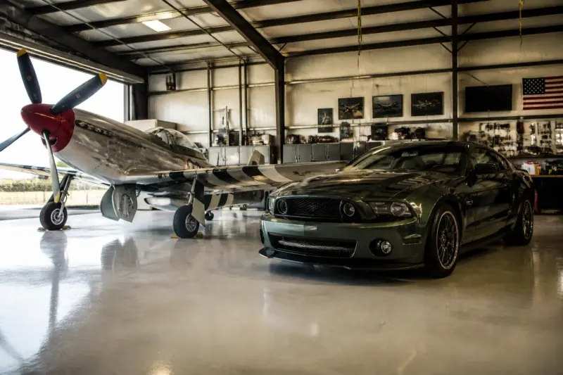 An American P-51 Mustang aircraft and a Roush Mustang car parked side by side in a hangar.